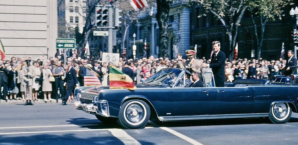 An open car flying Ethiopian and American flags passes crowds beside a Pennsylvania Avenue street sign