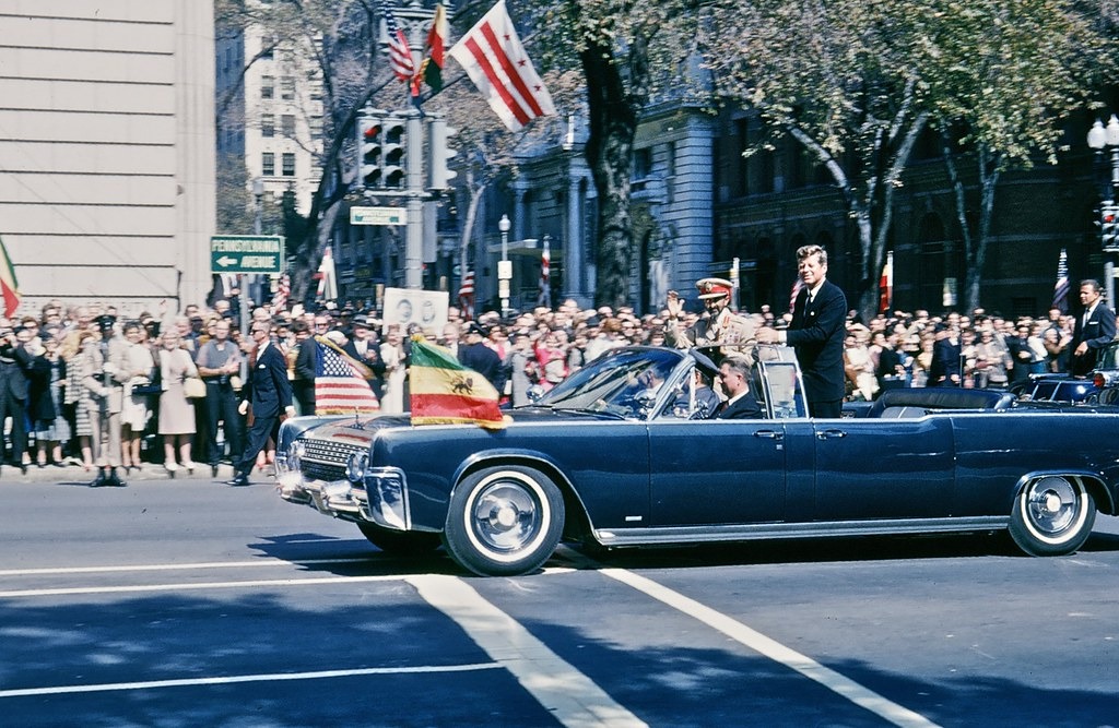An open car flying Ethiopian and American flags passes crowds beside a Pennsylvania Avenue street sign