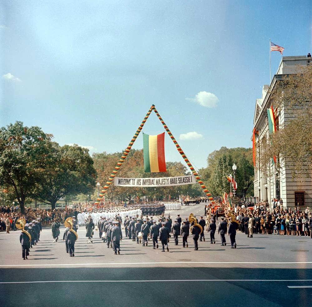 A military band marches past crowds beneath a banner reading Welcome to His Imperial Majesty Haile Selassie I and an Ethiopian flag